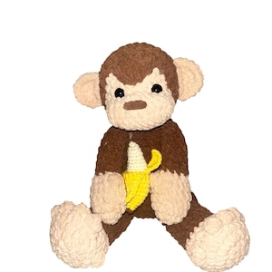 May include: A crocheted monkey plush toy with dark brown fur on the head, torso, and limbs, and light beige fur on the face, ears, hands, and feet. The monkey has black plastic eyes and holds a small, bright yellow crocheted banana in its hands.