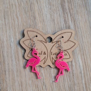 May include: A pair of bright pink flamingo earrings with silver-colored hooks. The earrings are displayed on a brown, butterfly-shaped tag with the text "Mad & Lolu Co."