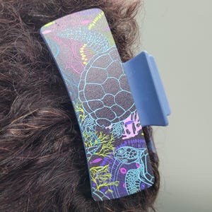 May include: A blue hair claw clip with a sea turtle design. The clip features a dark background with a colorful illustration of a sea turtle, coral, and other marine life. The clip is attached to dark curly hair.