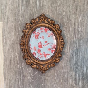 May include: Oval-shaped wooden mirror with ornate detailing. The mirror's surface is light blue with red handprints and streaks. The frame is a dark brown color, and the mirror is set against a gray wooden surface.