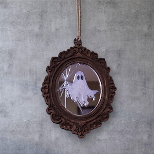 May include: A Halloween-themed decorative mirror with a brown, ornate frame. The mirror features a ghostly figure and a spiderweb design. A natural twine loop is attached for hanging. The mirror is a unique piece of seasonal decor.