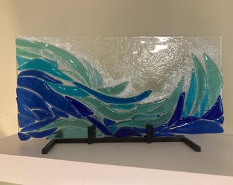 Könnte beinhalten: Ein Glaskunstwerk, das eine Welle mit blauen und grünen Glasstücken darstellt. Die Welle befindet sich auf einem klaren Glasgrund und ist auf einem schwarzen Metallständer montiert.