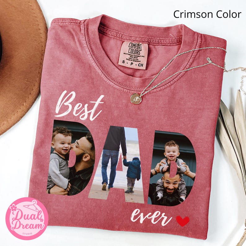 May include: Crimson-coloured t-shirt with the words "Best Dad Ever" and a heart graphic. The word "DAD" features family photos. A silver necklace with a pendant is on the shirt.
