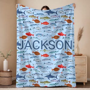 May include: A light blue blanket featuring a pattern of various fish in shades of red, brown, blue, and green. The name "JACKSON" is printed in large, blue letters across the blanket. The blanket is held up by a person.