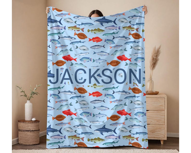 May include: A light blue blanket featuring a pattern of various fish in shades of red, brown, blue, and green. The name "JACKSON" is printed in large, blue letters across the blanket. The blanket is held up by a person.