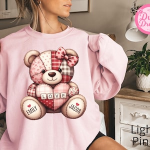 May include: Pale pink sweatshirt with a patchwork teddy bear design. The bear has a heart-shaped chest with the word "LOVE" and the names "Emily" and "Jacob". The bear wears a bow and has a mix of patterns.