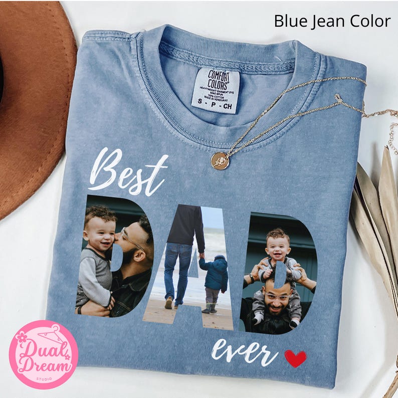 May include: Blue jean coloured t-shirt with the words "Best Dad Ever" in white script. The word "DAD" features family photos. A gold necklace with a pendant is draped across the shirt. The shirt is a Comfort Colors brand.