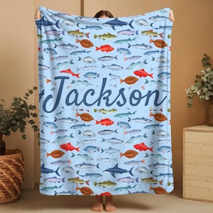 May include: A light blue blanket featuring a pattern of various fish in shades of red, brown, gray, and blue. The name "Jackson" is written in a cursive font across the center of the blanket. The blanket is held up by a person.