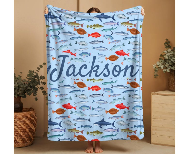 May include: A light blue blanket featuring a pattern of various fish in shades of red, brown, gray, and blue. The name "Jackson" is written in a cursive font across the center of the blanket. The blanket is held up by a person.