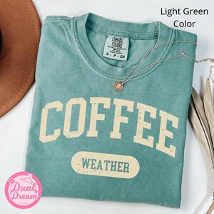 K&ouml;nnte beinhalten: Hellgr&uuml;nes T-Shirt mit dem Text "COFFEE WEATHER" in einer Vintage-Schriftart. Das Shirt ist aus weichem, bequemem Material und hat eine lockere Passform.
