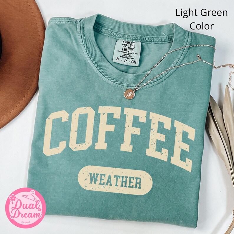 K&ouml;nnte beinhalten: Hellgr&uuml;nes T-Shirt mit dem Text "COFFEE WEATHER" in einer Vintage-Schriftart. Das Shirt ist aus weichem, bequemem Material und hat eine lockere Passform.