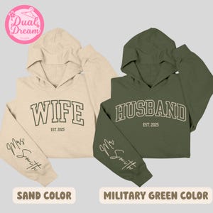 May include: Two hooded sweatshirts, one in sand color and one in military green, with the words "Wife" and "Husband" printed on the front. The text "Est. 2025" is printed below the words. The sleeves of both sweatshirts have the text "Mrs. Smith" written in cursive.