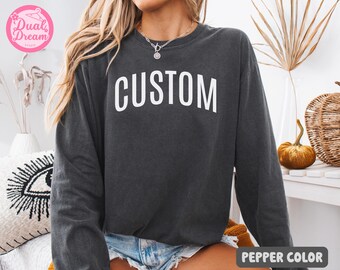 May include: A dark gray sweatshirt with the word "CUSTOM" in white block letters. The long-sleeved top is a pepper color and has a relaxed fit. The sweatshirt is a casual style.
