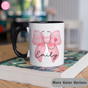 May include: White ceramic mug with a black rim and handle. The mug features a pink bow with a stethoscope and the name "Emily" in black cursive.