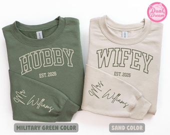 May include: A military green sweatshirt with the text HUBBY EST. 2026 and Mr. Williams on the sleeve, and a sand-colored sweatshirt with the text WIFEY EST. 2026 and Mrs. Williams on the sleeve. Both feature crew necks and long sleeves.
