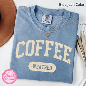 K&ouml;nnte beinhalten: Ein T-Shirt in Jeansblau mit dem Text "COFFEE WEATHER" in einer Vintage-Schriftart. Das Shirt ist aus einem weichen, bequemen Material.