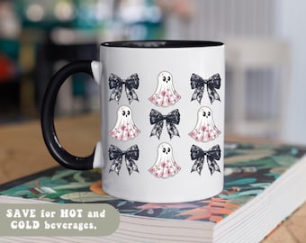 May include: A white ceramic mug with a black handle and interior rim. The exterior features a repeating pattern of black lace-style bows and white ghost figures decorated with small pink floral designs. The text "SAVE for HOT and COLD beverages."