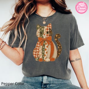 Puede incluir: Una camiseta de manga corta gris carbón con un gráfico central de un gato compuesto por varios patrones de patchwork en naranja, verde y blanco, incluyendo calabazas, flores y cuadros, con un gran lazo naranja en el cuello del gato.