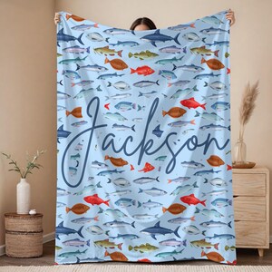 May include: A light blue blanket featuring a pattern of various fish in shades of red, brown, blue, and green. The name "Jackson" is written in a cursive font across the blanket. The blanket is held up by a person.