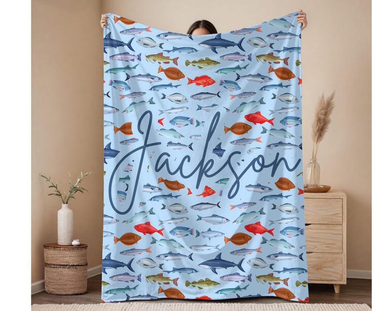 May include: A light blue blanket featuring a pattern of various fish in shades of red, brown, blue, and green. The name "Jackson" is written in a cursive font across the blanket. The blanket is held up by a person.