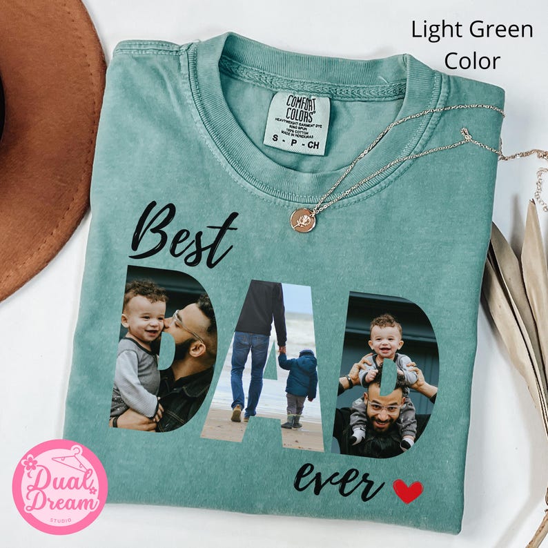 May include: Light green t-shirt with the words "Best Dad Ever" in black script. The letters of "DAD" are cut out to reveal three photos. A silver necklace with a pendant is on the shirt.