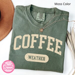 K&ouml;nnte beinhalten: Ein gr&uuml;nes T-Shirt mit dem Text "COFFEE WEATHER" in einer Vintage-Schriftart. Das Shirt ist aus weichem, bequemem Material und hat eine lockere Passform.