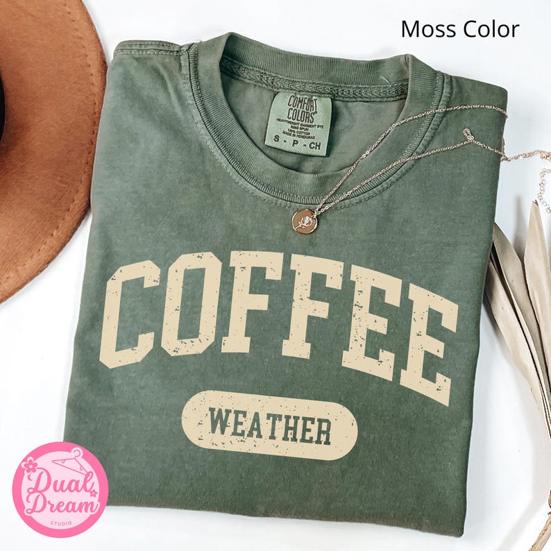 K&ouml;nnte beinhalten: Ein gr&uuml;nes T-Shirt mit dem Text "COFFEE WEATHER" in einer Vintage-Schriftart. Das Shirt ist aus weichem, bequemem Material und hat eine lockere Passform.