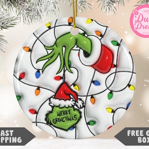 May include: A round Christmas ornament featuring the Grinch's hand and face, with a red Santa hat and the words "Merry Grinchmas." The ornament is decorated with colorful Christmas lights and a gold string for hanging.
