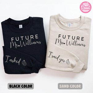 Può includere: Due felpe, una nera e una color sabbia, con il testo "FUTURE Mr. Williams" e "FUTURE Mrs. Williams" su di esse. La felpa nera ha il testo "I asked" sulla manica e la felpa color sabbia ha il testo "I said yes" sulla manica.