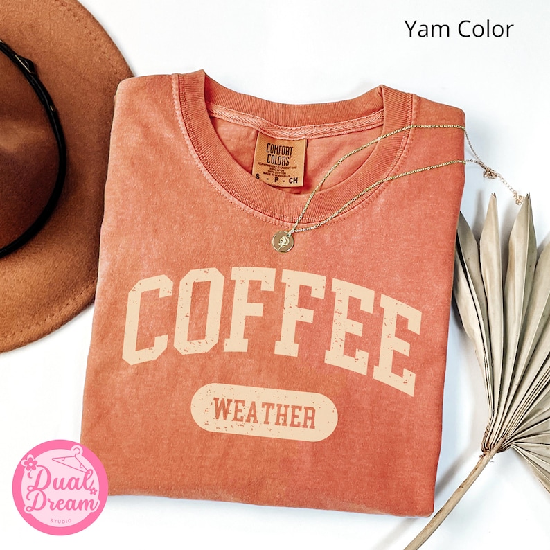 K&ouml;nnte beinhalten: Ein verbrannt-orangenes T-Shirt mit dem Text "COFFEE WEATHER" in einer Vintage-Universit&auml;ts-Schriftart. Das Shirt ist aus einem weichen, bequemen Material und hat einen Used-Look.