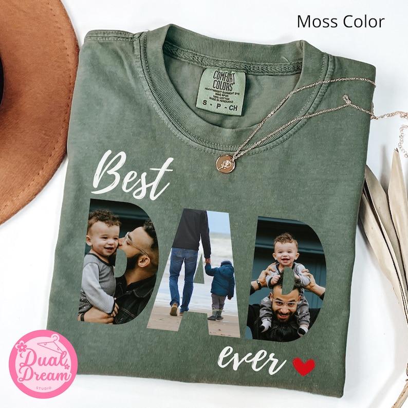 May include: A moss-green t-shirt with the words "Best Dad Ever" in white script. The word "DAD" features three family photos. A gold necklace with a pendant is draped on the shirt. A red heart graphic is below the word "ever".
