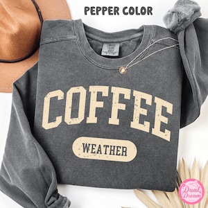 May include: A gray sweatshirt with the text "COFFEE WEATHER" in a distressed, vintage-style font. The sweatshirt is a comfortable, casual style.