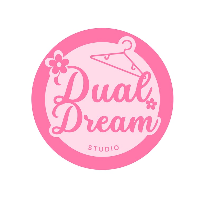 Op de afbeelding: Roze cirkelvormig logo met de tekst "Dual Dream Studio" in een wit cursief lettertype. Een witte kleerhanger met een haak bevindt zich boven de tekst. Een kleine witte bloem bevindt zich links van de tekst en een andere kleine witte bloem bevindt zich rechts van de tekst.