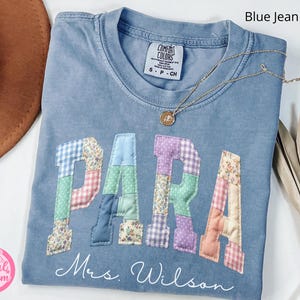May include: Blue jean colored t-shirt with the word "PARA" in patchwork letters. The name "Mrs. Wilson" is written below the letters. A gold necklace with a pendant is draped across the shirt.