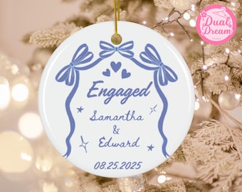 May include: White ceramic ornament with a blue design and gold string. The ornament features the word "Engaged", names "Samantha & Edward", and the date "08.25.2025". Decorative bows, hearts, and stars complete the design.