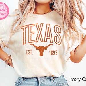 May include: Ivory-colored t-shirt with the word "TEXAS" in distressed orange lettering. Below the word "TEXAS" is a longhorn graphic and the text "EST. 1893". The shirt is a crew neck style.