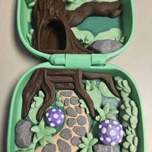 Puede incluir: Un juguete verde en forma de concha, impreso en 3D y abierto, con una escena de bosque. La escena incluye un &aacute;rbol marr&oacute;n, un camino de piedra, setas moradas y blancas y follaje verde. El juguete est&aacute; dise&ntilde;ado para abrirse y cerrarse.