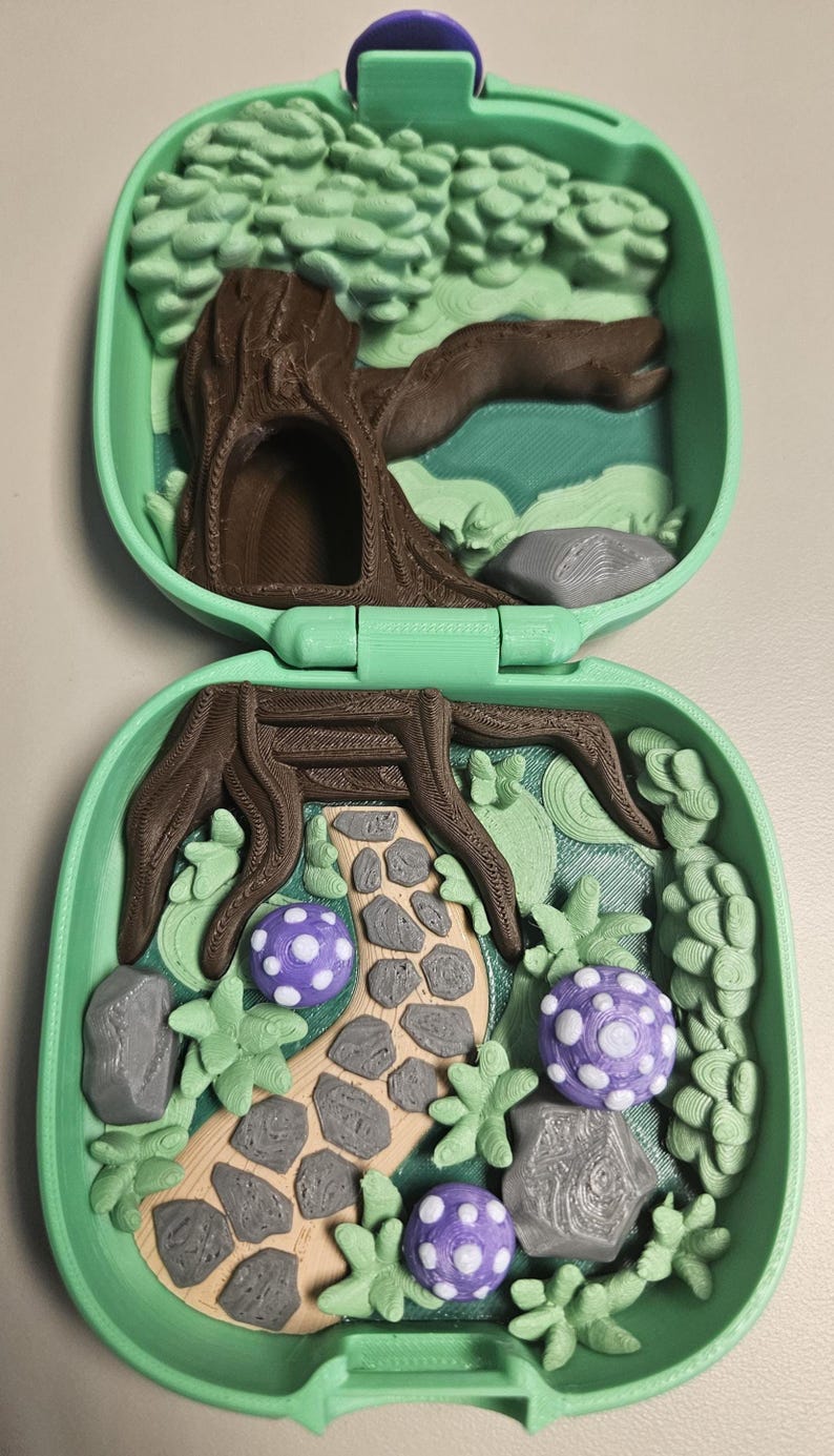 Puede incluir: Un juguete verde en forma de concha, impreso en 3D y abierto, con una escena de bosque. La escena incluye un &aacute;rbol marr&oacute;n, un camino de piedra, setas moradas y blancas y follaje verde. El juguete est&aacute; dise&ntilde;ado para abrirse y cerrarse.