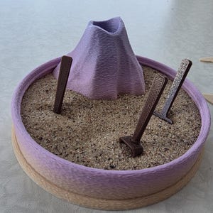 May include: A miniature zen garden with a lavender volcano, brown rakes, and sand. The round base has a gradient from tan to lavender. The volcano has a textured surface. The rakes are angled in the sand.