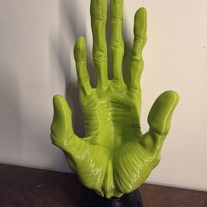 May include: A lime green, open-palmed hand sculpture. The hand is mounted on a black, circular base. The fingers are long and slightly curved. The sculpture is made of a smooth, glossy material.
