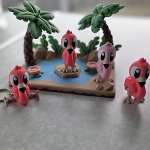 May include: A miniature diorama featuring a tropical scene with cartoon flamingos. The scene includes a blue pond, palm trees, and pink flowers. Several pink flamingo figurines are positioned around the scene, some with keychains.
