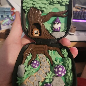 Puede incluir: Una escena de bosque en miniatura dentro de una caja negra. La escena incluye un &aacute;rbol con una criatura en un hueco, un mago, un camino de piedra y setas moradas con lunares. Los colores son verde, marr&oacute;n, gris y morado.