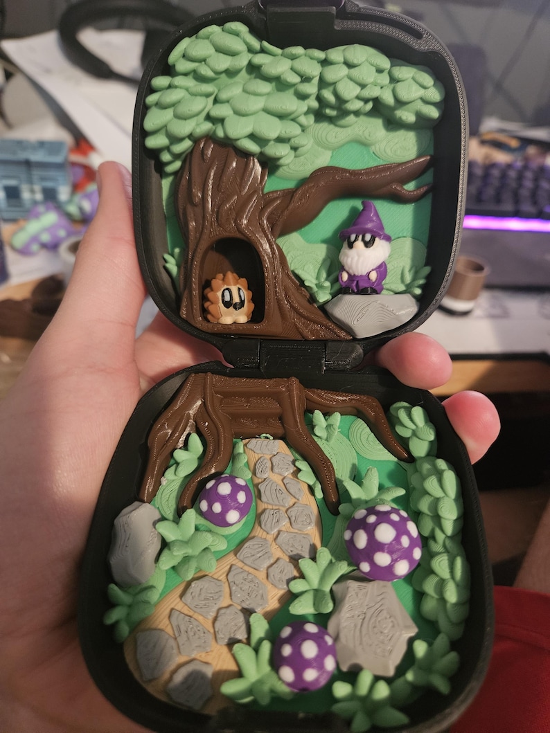 Puede incluir: Una escena de bosque en miniatura dentro de una caja negra. La escena incluye un &aacute;rbol con una criatura en un hueco, un mago, un camino de piedra y setas moradas con lunares. Los colores son verde, marr&oacute;n, gris y morado.