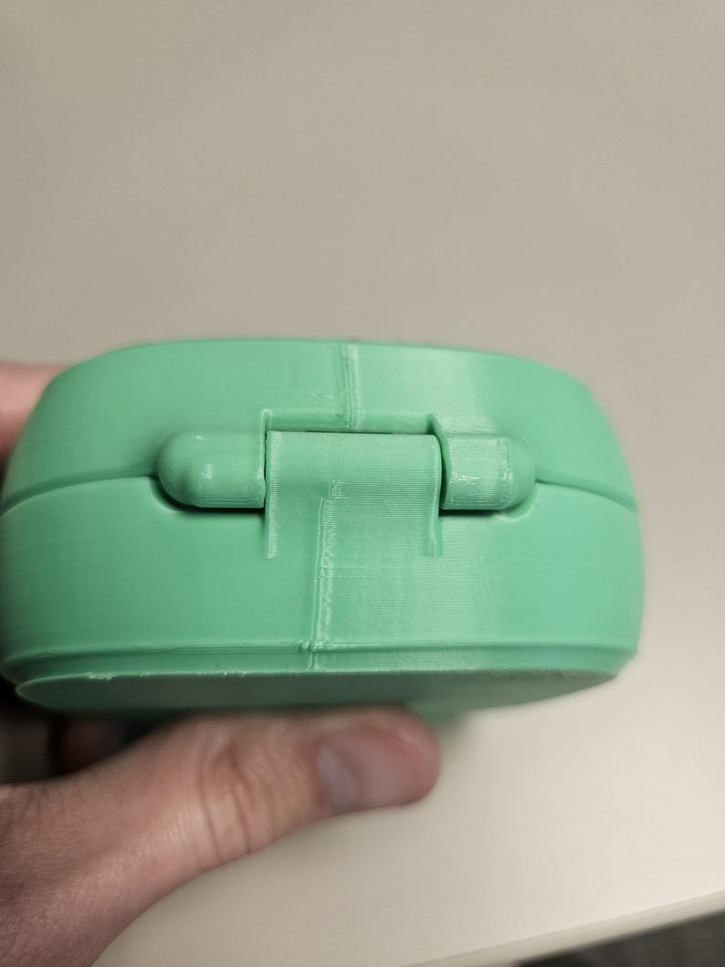 Puede incluir: Un recipiente verde claro, redondeado y con bisagras. El recipiente est&aacute; hecho de un material s&oacute;lido y tiene una bisagra visible. El recipiente est&aacute; sostenido por una mano. El recipiente es de color verde claro liso.