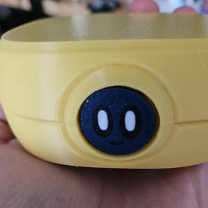 Puede incluir: Primer plano de un objeto amarillo y redondeado con un bot&oacute;n circular azul oscuro. El bot&oacute;n presenta una cara de dibujos animados con dos ojos ovalados blancos y una peque&ntilde;a boca curvada. El objeto parece ser de pl&aacute;stico.