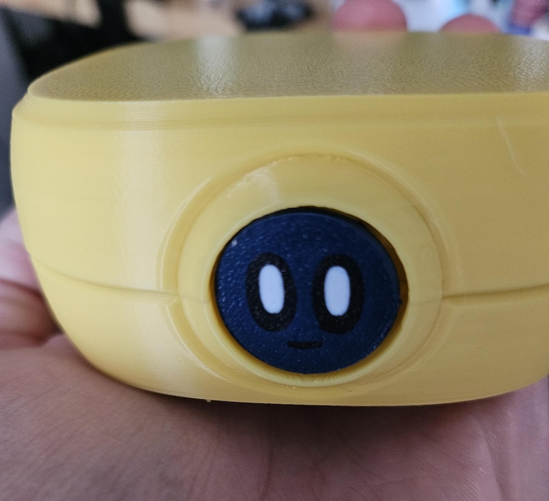 Puede incluir: Primer plano de un objeto amarillo y redondeado con un bot&oacute;n circular azul oscuro. El bot&oacute;n presenta una cara de dibujos animados con dos ojos ovalados blancos y una peque&ntilde;a boca curvada. El objeto parece ser de pl&aacute;stico.