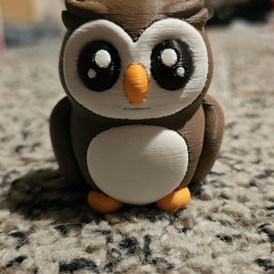 May include: A brown and white 3D printed owl figurine with large eyes and an orange beak. The owl is standing on a brown carpet.