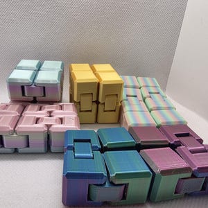 May include: A collection of colorful fidget toys in various shades, including pink, yellow, blue, and rainbow hues. The toys are made of connected blocks that can be manipulated and moved. The background is a plain white surface.