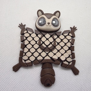 May include: A 3D-printed, brown and tan flying squirrel figurine. The squirrel has large, detailed eyes, small ears, and outstretched wings with a patterned design. The tail is segmented, and the figure is set against a white background.