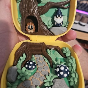 Puede incluir: Un juguete amarillo, abierto y port&aacute;til con una escena de bosque en miniatura. La escena incluye un &aacute;rbol con una criatura en un hueco, un gnomo, setas y un camino de piedra. El juguete es de pl&aacute;stico y tiene un dise&ntilde;o 3D detallado.